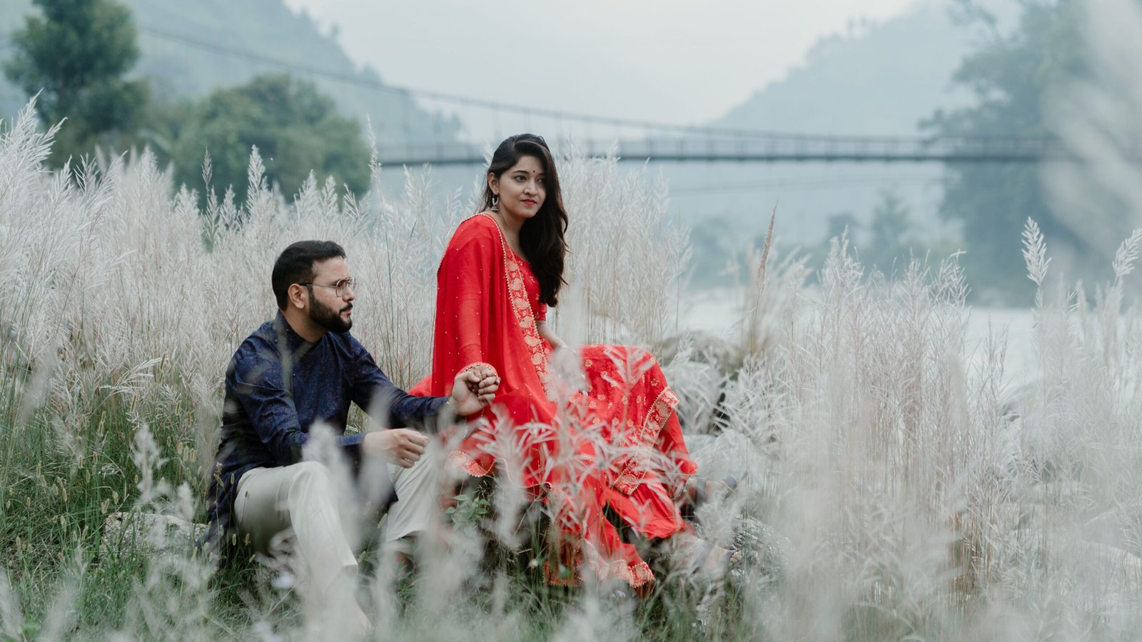 A couple in traditional attire sitting in a serene grass field with a scenic background.