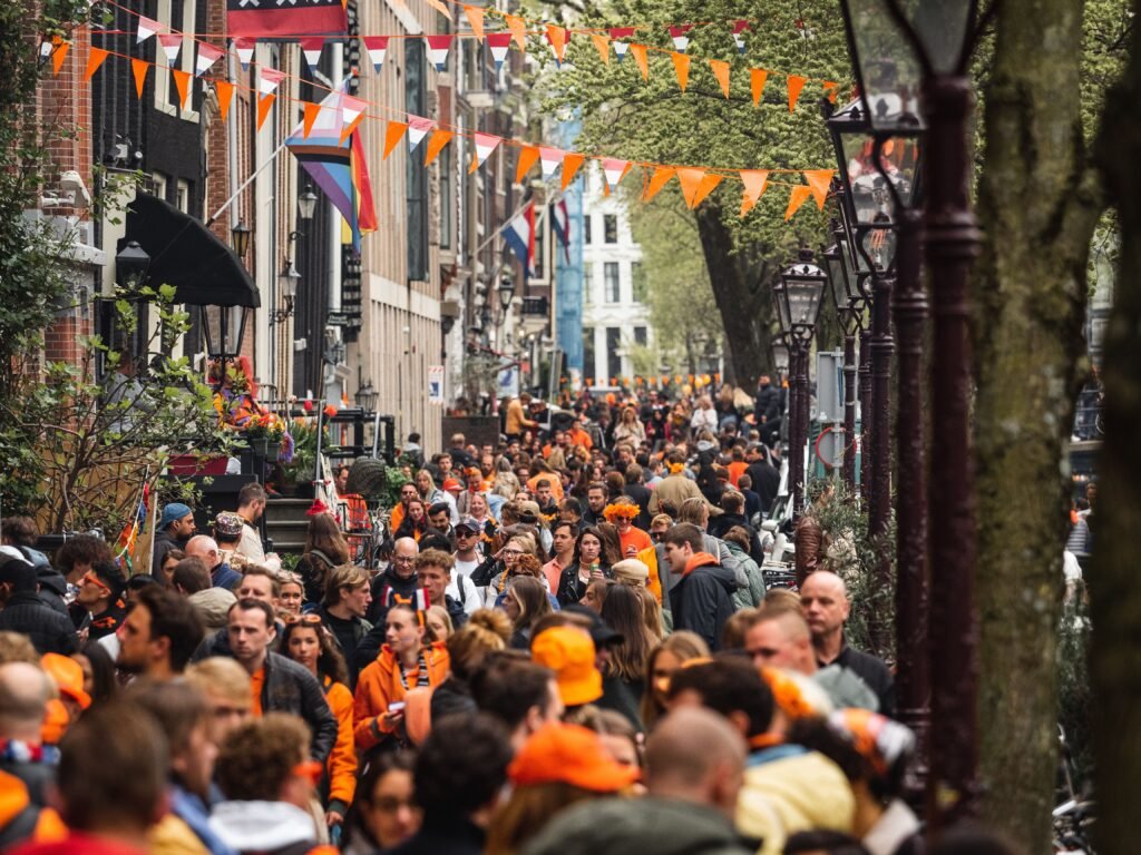 A vibrant street festival with a large, diverse crowd celebrating in a city setting with colorful flags.
